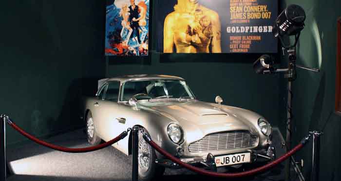 Classic silver car displayed at an exhibition with movie posters and camera, featured on digipdemo.com's Iconic Movie Cars article.